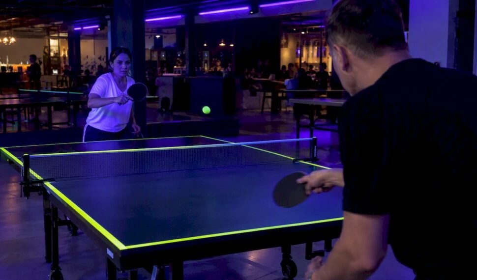 Players rally beneath blacklights at a ping pong table